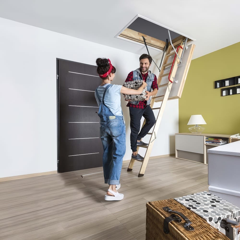 A Man And Woman Are Happily Passing A Box To Each Other As The Man Climbs A Wooden Attic Ladder In A Modern Well-Lit Room
