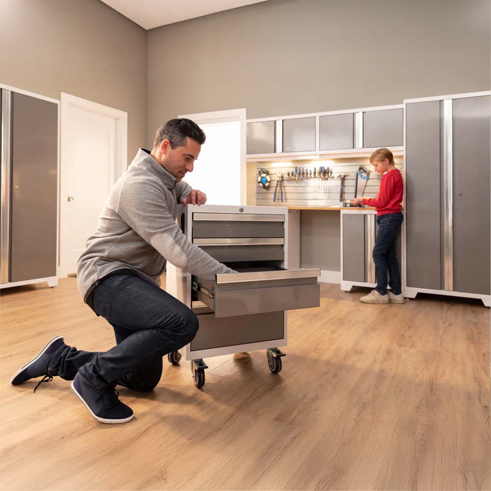A Man Kneeling To Organize Tools In A Mobile Drawer Cabinet While A Child Works At A Nearby Workstation