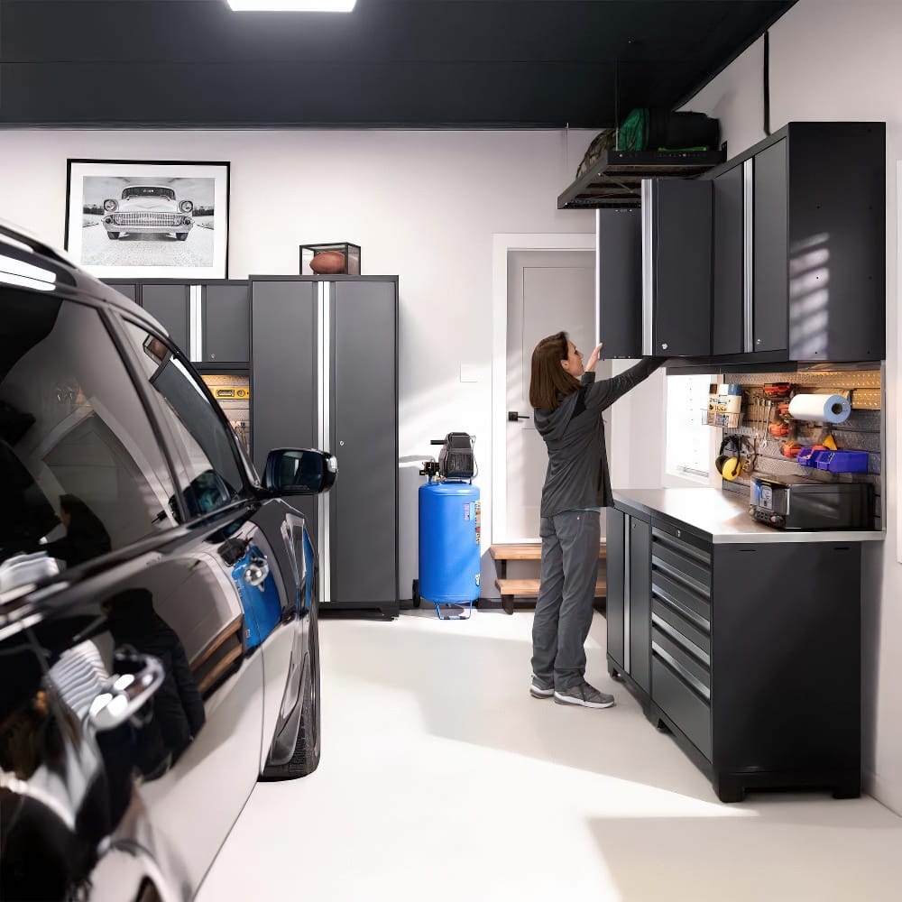 A Woman Reaches Into A Wall-Mounted Garage Cabinet Beside A Tool-Filled Workstation With A Car Parked Nearby