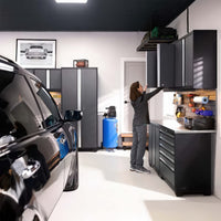 A Woman Reaches Into A Wall-Mounted Garage Cabinet Beside A Tool-Filled Workstation With A Car Parked Nearby