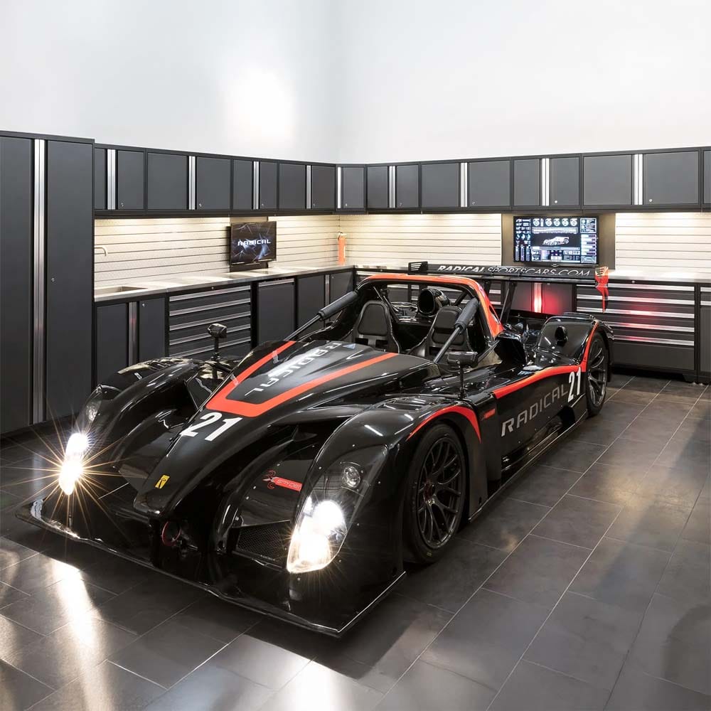 Black And Red Race Car With Lights Shining Parked In High End Garage Featuring Long Wall Of Dark Cabinets And Tool Drawers