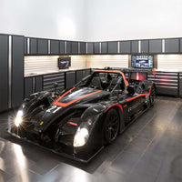 Black And Red Race Car With Lights Shining Parked In High End Garage Featuring Long Wall Of Dark Cabinets And Tool Drawers