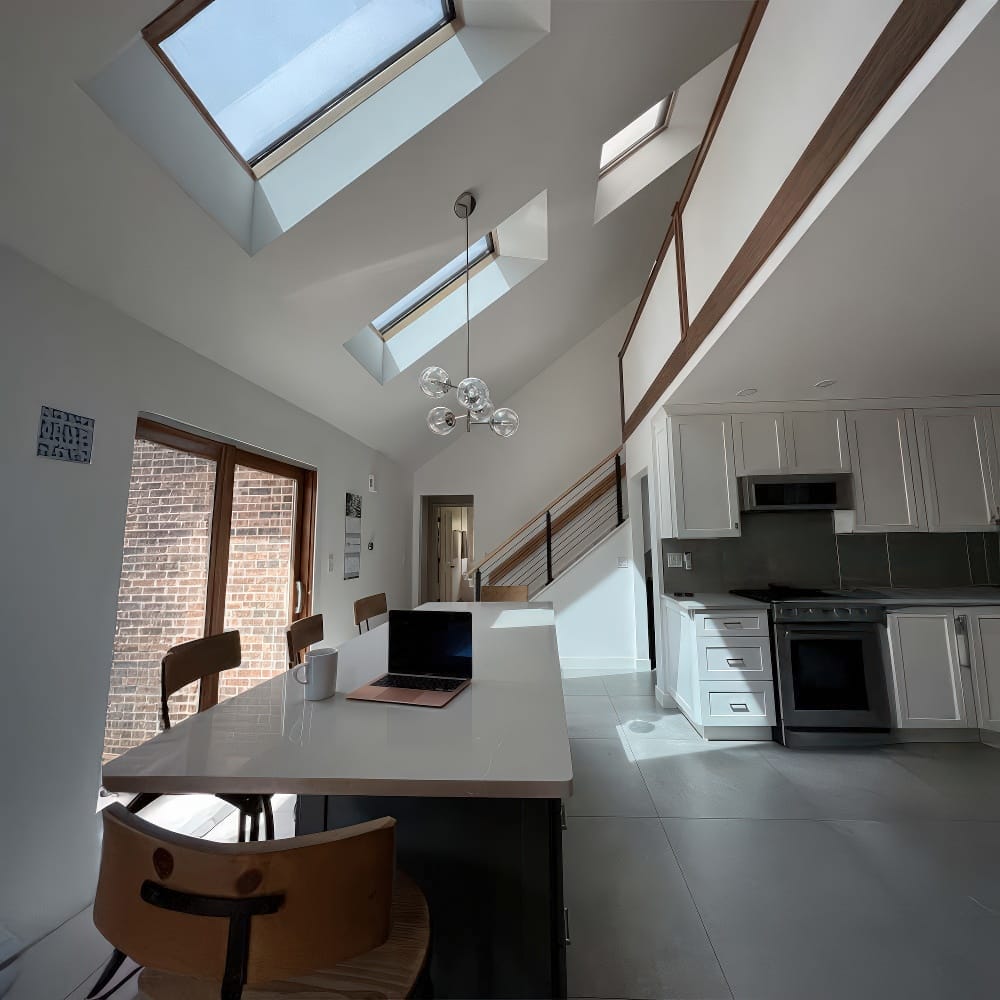 Bright Kitchen And Dining Area With Skylights A Long White Island A Laptop On The Table And Modern Wood Accents