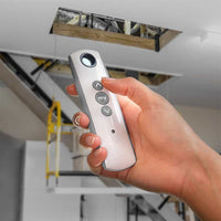 Hand holding a remote control for operating an electric attic ladder with open ceiling access.