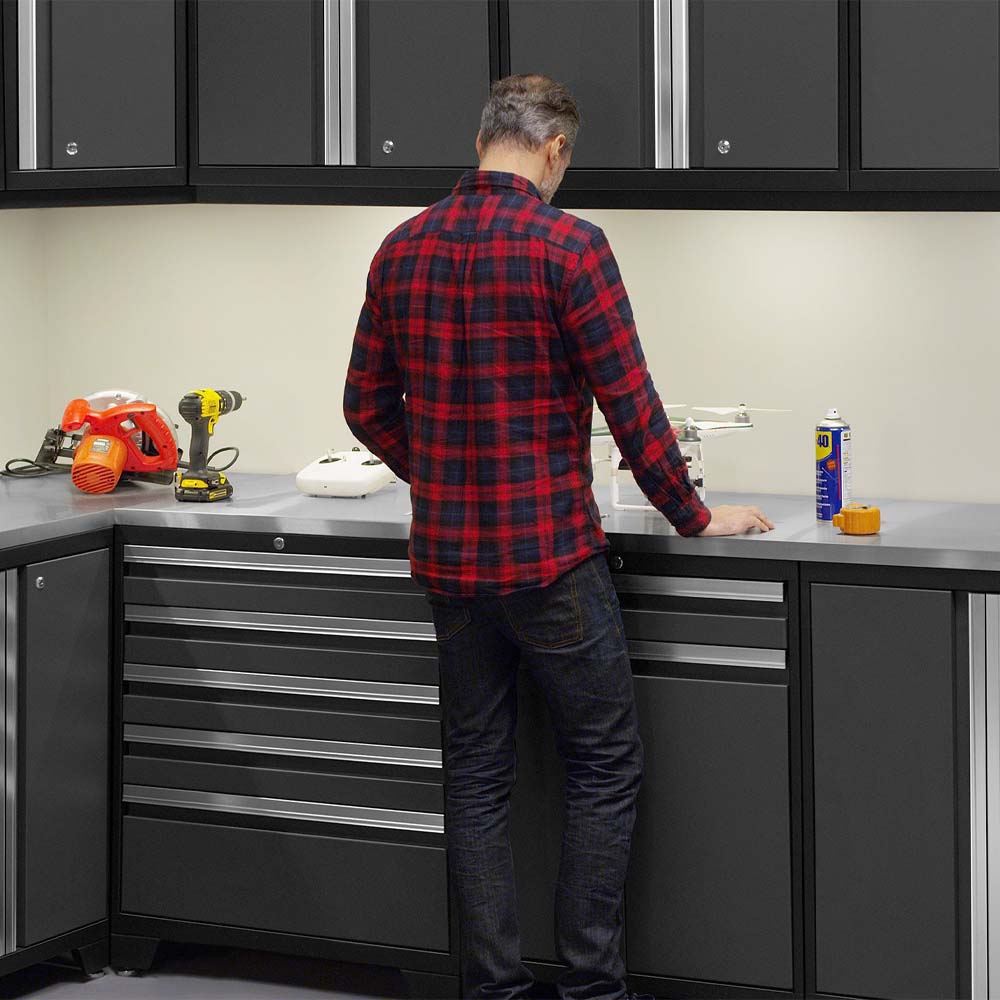 Man In A Red Plaid Shirt Stands At A Garage Workbench Surrounded By Dark Cabinets And Assorted Power Tools