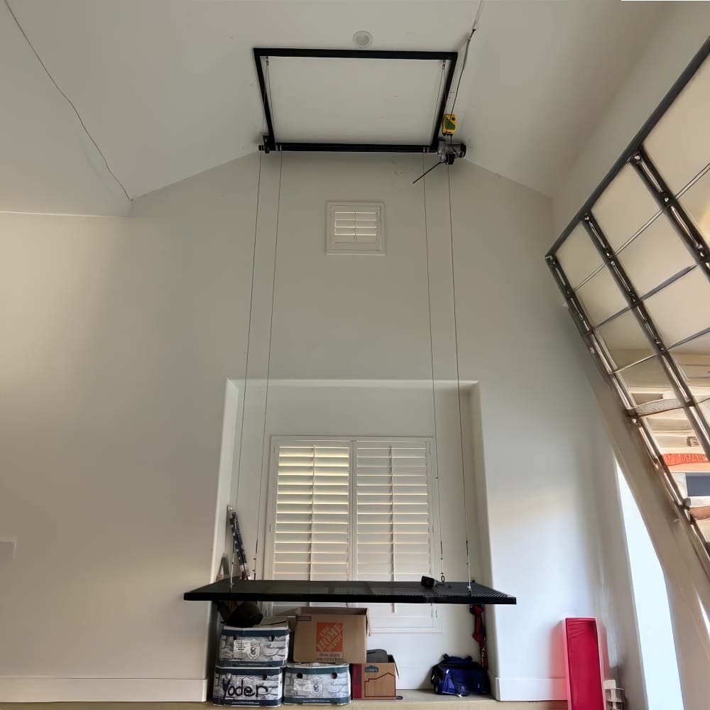 Motorized Ceiling Hoist Lowers Mesh Storage Platform On Cables In Tall Garage With Window Shutters And Boxes Below