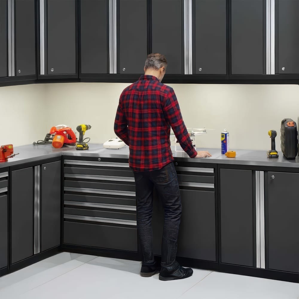 Person In Plaid Shirt Leans Over Corner Workbench Surrounded By Matte Gray Cabinets And Assorted Power Tools