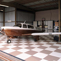 Small Aircraft With White And Brown Exterior Parked Inside A Hangar With A Checkered Racedeck Flooring
