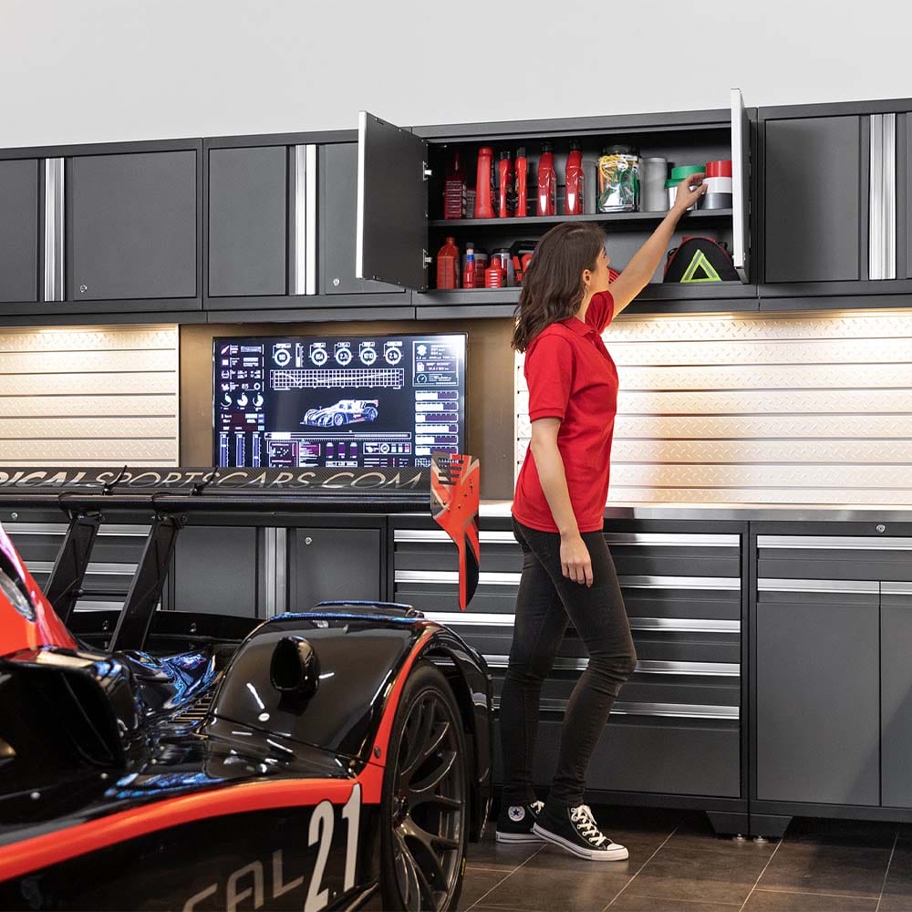 Woman In Red Shirt Reaching Into Overhead Garage Cabinet Above Race Car Workstation With Monitor Display