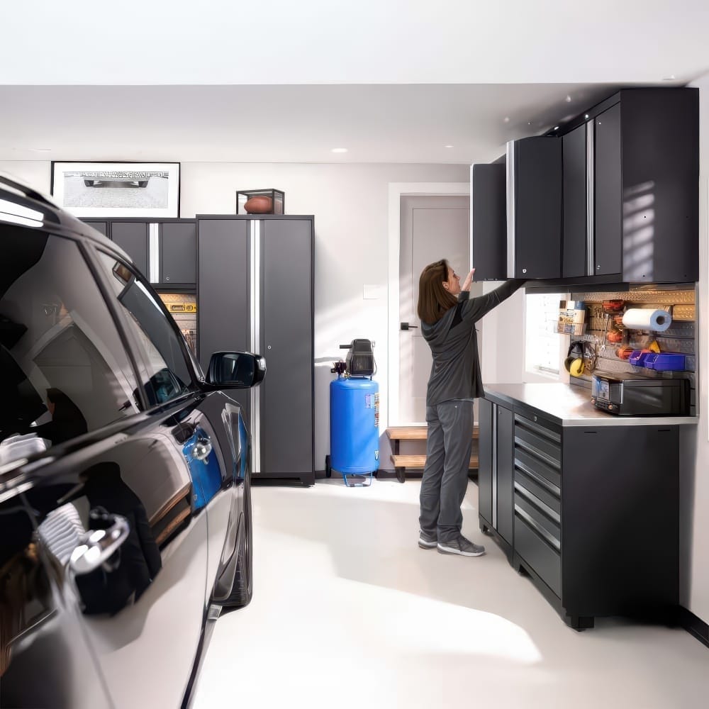 Woman Reaches Garage Wall Cabinets In Organized Workspace With Black Car Blue Air Compressor And Tool Bench