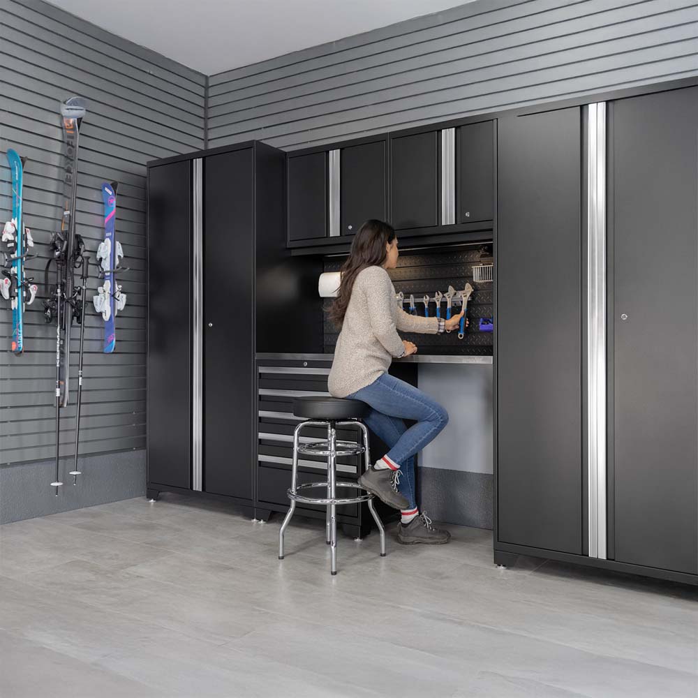 Woman Sitting On Stool In Modern Garage Workspace Surrounded By Black Cabinets And Wall Mounted Skis